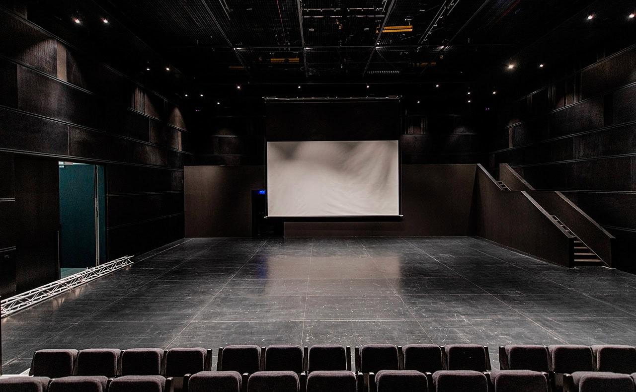 Black box theatre with a large white projection screen, dark walls and flexible floor seating.