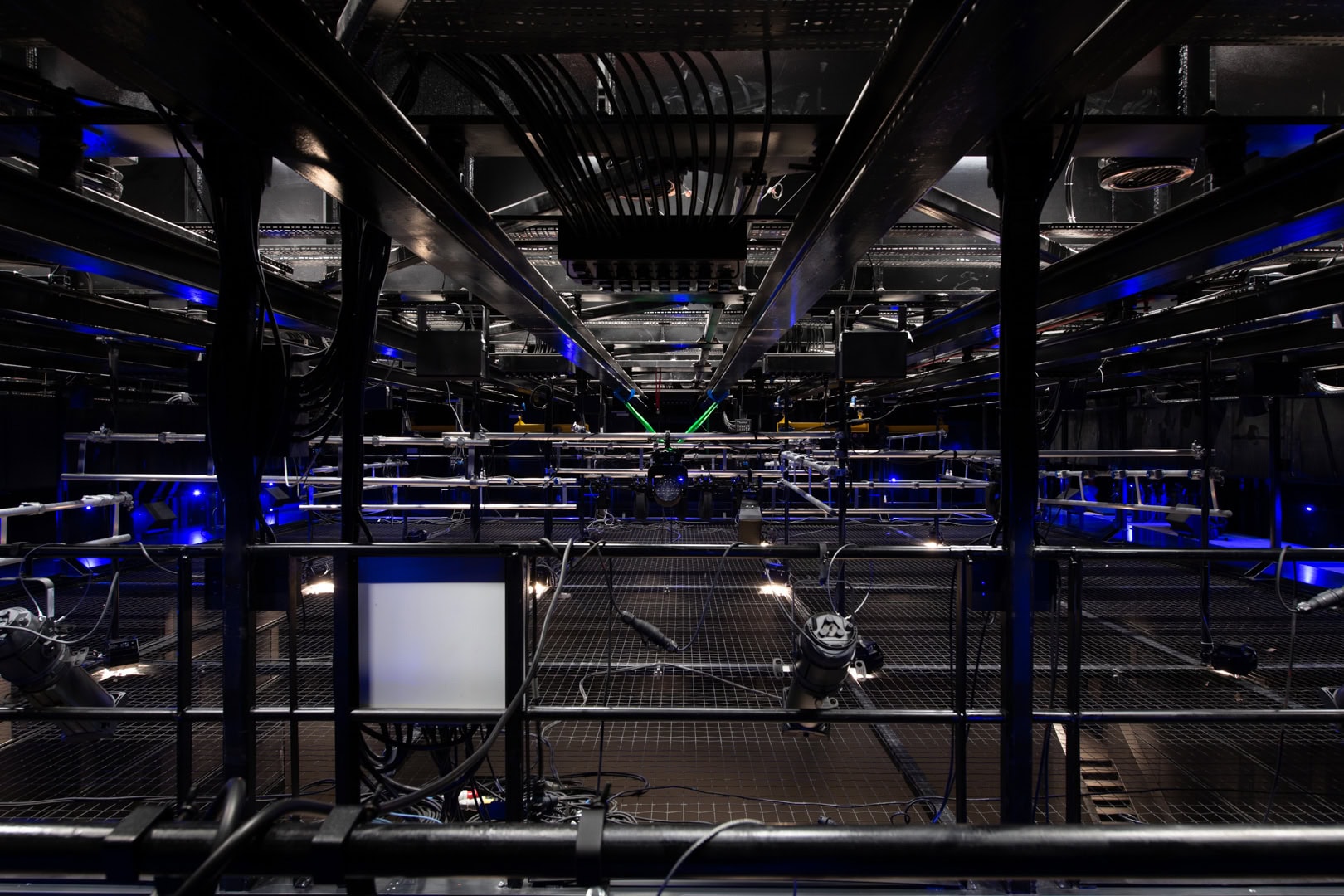 Technical gallery above a black box theatre, exposed lighting rig and structural grid under blue illumination.