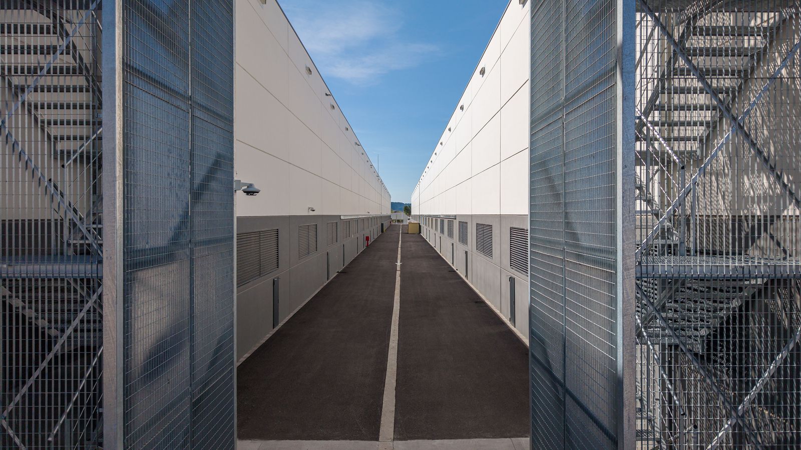 Aerial view straight down a narrow service corridor between two warehouse blocks, external stairs and mesh screens on both sides.