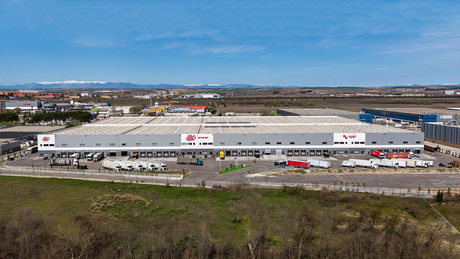 Aerial overview of a large logistics warehouse and its lorry yard in the Madrid industrial corridor, open country beyond.