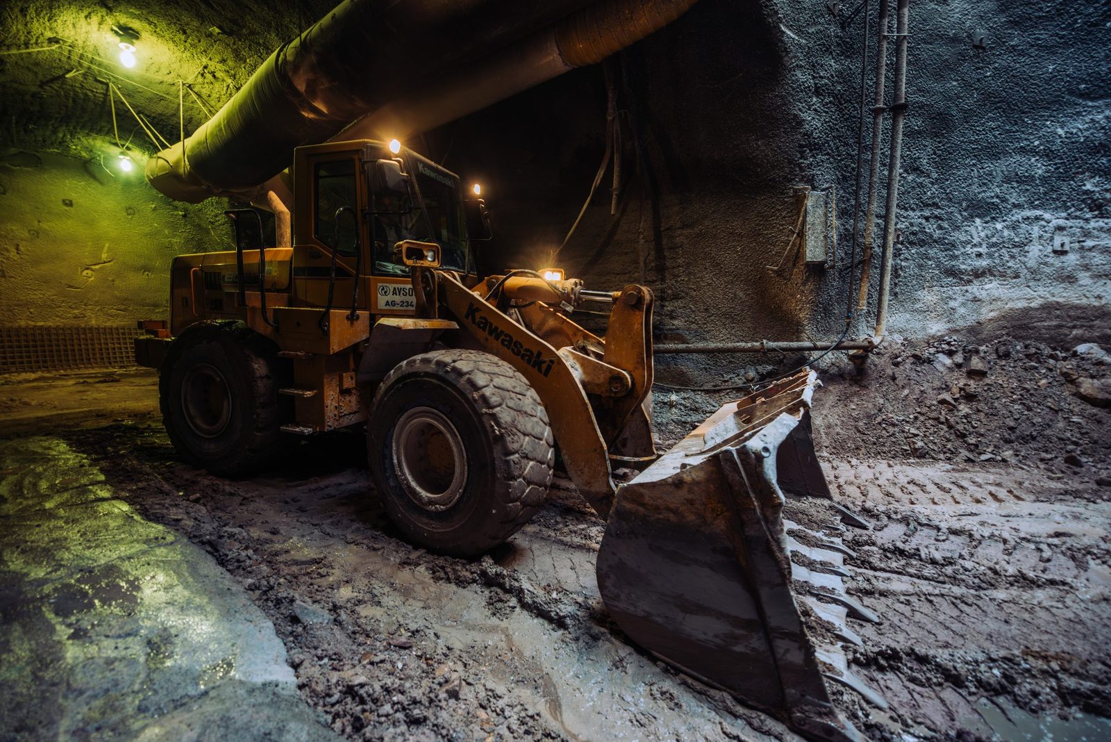 Aerial view of underground construction site with yellow bulldozer working inside a tunnel, illuminated with green and yellow lighting, showing rough concrete walls, pipes, and construction debris.