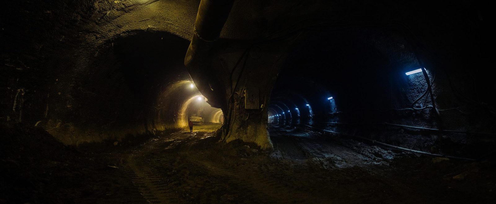 Underground tunnel construction at night with artificial lighting, showing a worker in the distance and the rugged interior walls of the tunnel.