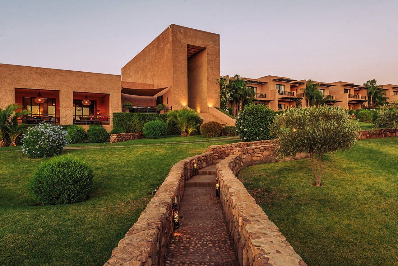 Resort marroquí al anochecer, arquitectura en tonos tierra con un camino empedrado que asciende entre céspedes y olivos.