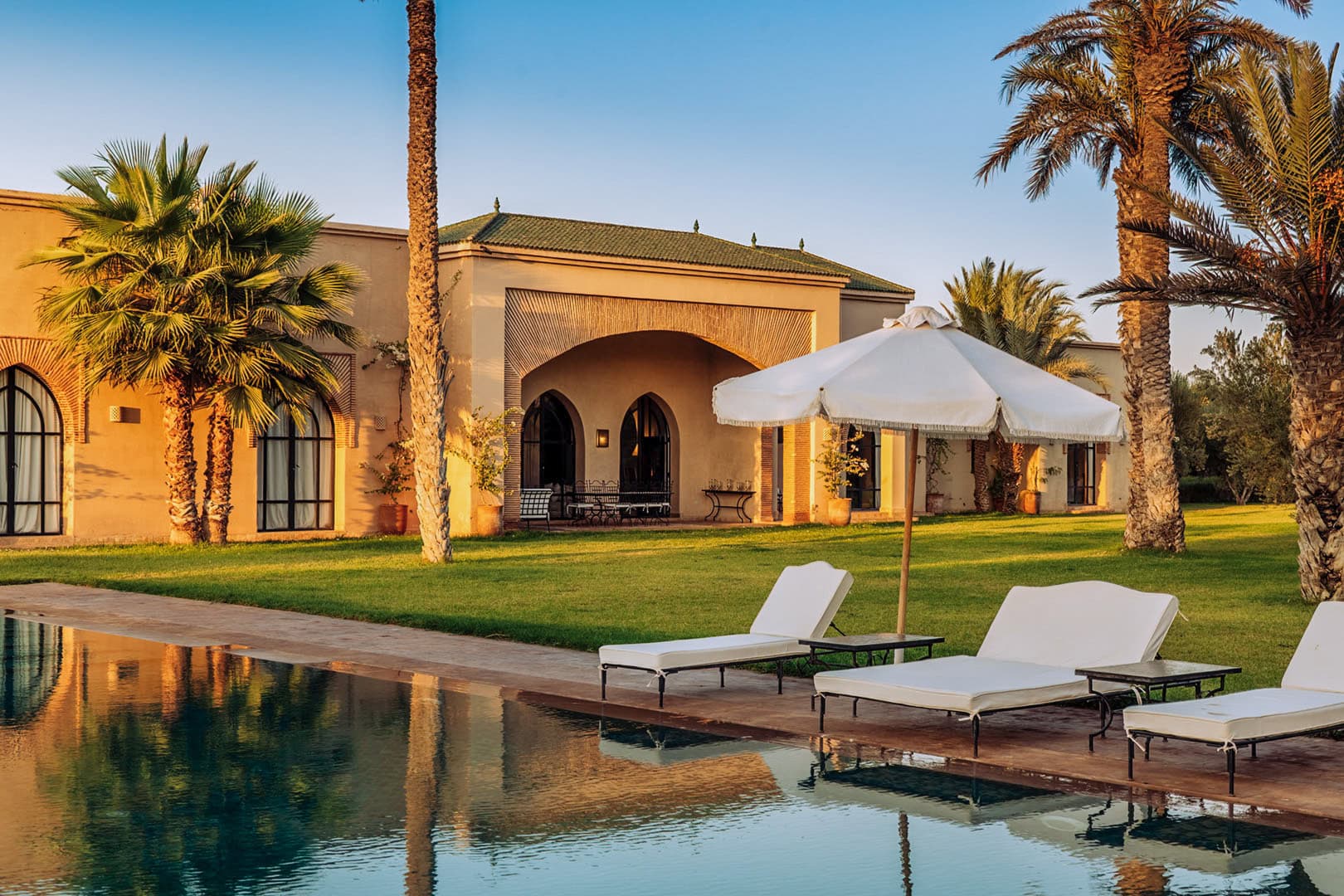 Exterior de villa marroquí de lujo con piscina durante la hora dorada, con palmeras y mobiliario exterior premium para marketing de hostelería de alto nivel