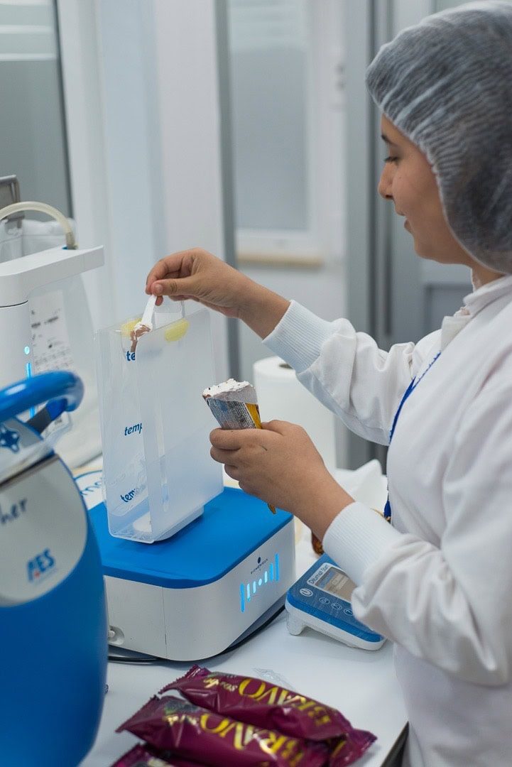 Quality control technician in hairnet and lab coat inspecting a product sample in a food safety laboratory.