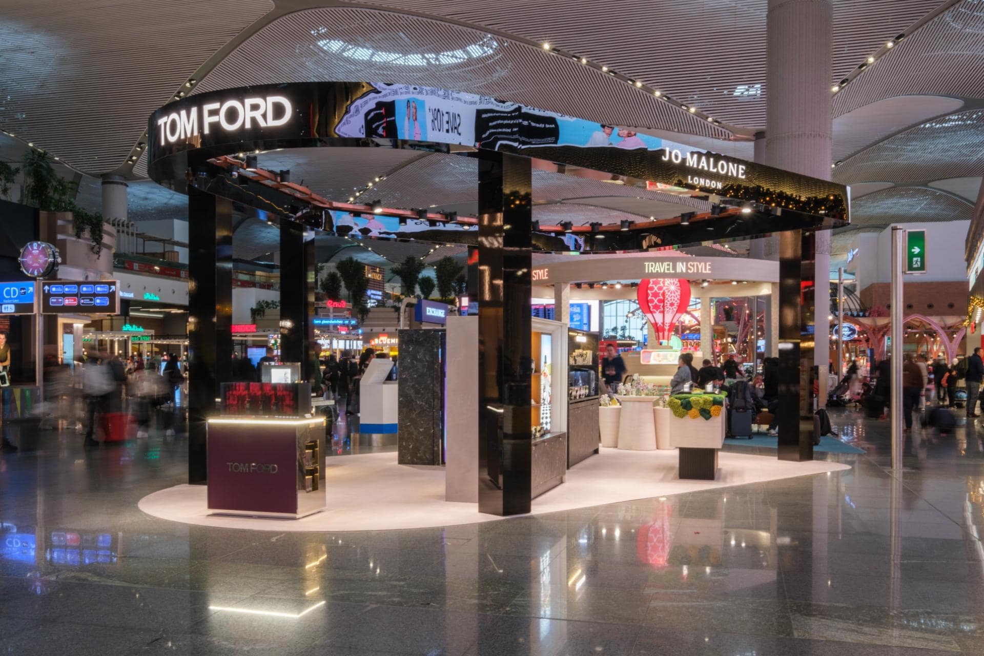 Tom Ford and Jo Malone retail kiosk in a busy airport terminal with passengers passing.