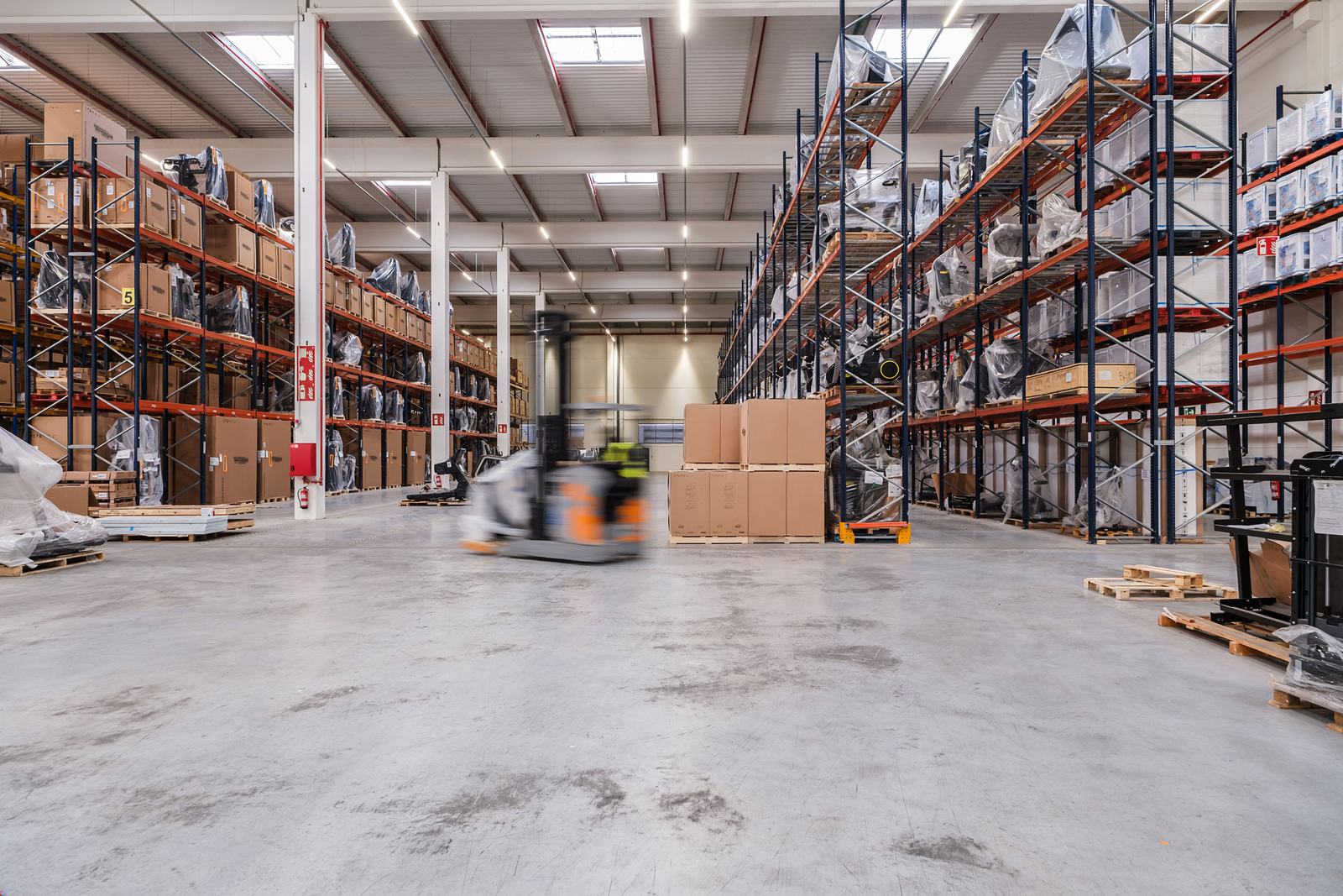 Warehouse floor with a forklift crossing between racking runs, motion visible against a stack of cartons.
