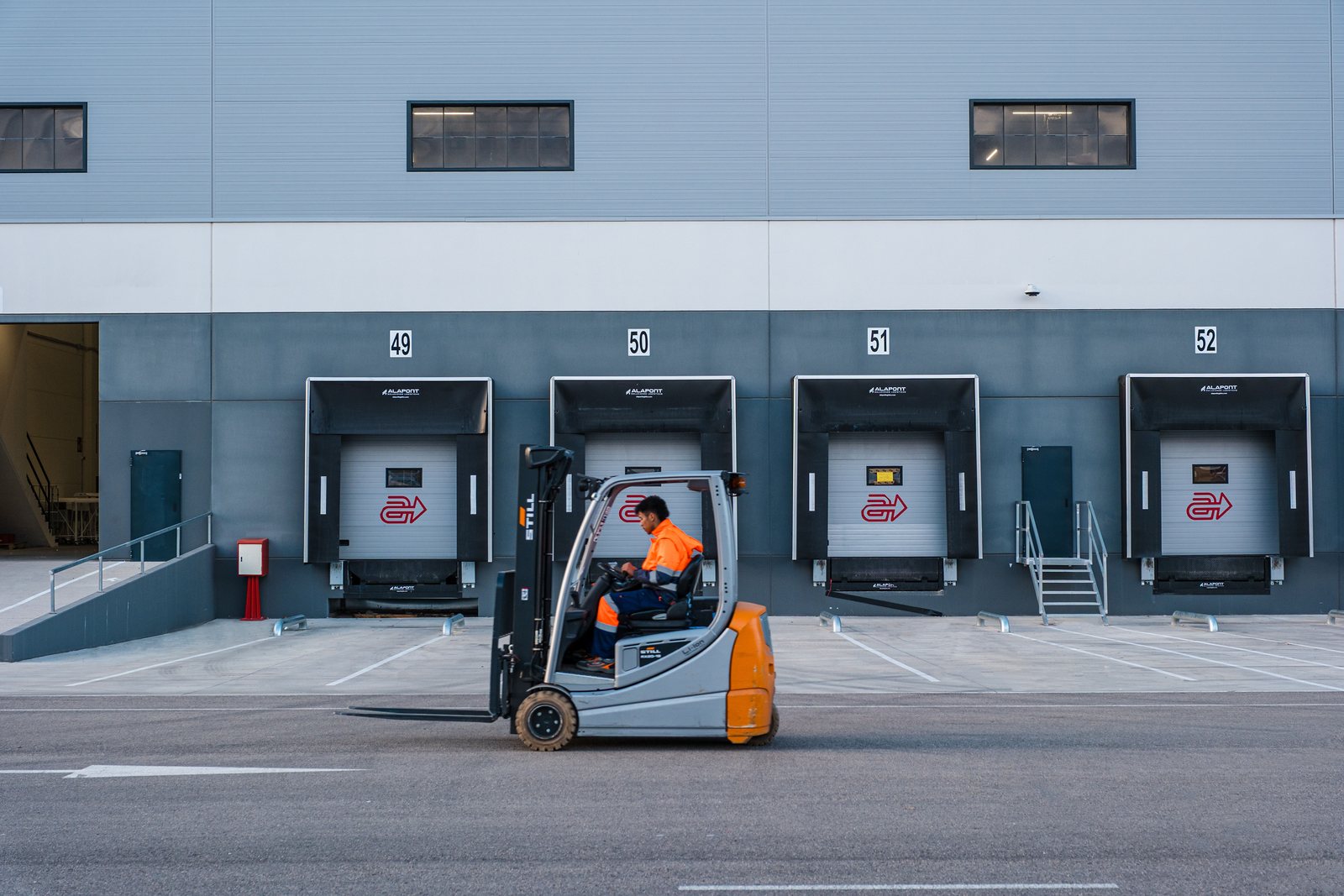Numbered loading dock doors along a grey warehouse elevation with a forklift and operator crossing in front.