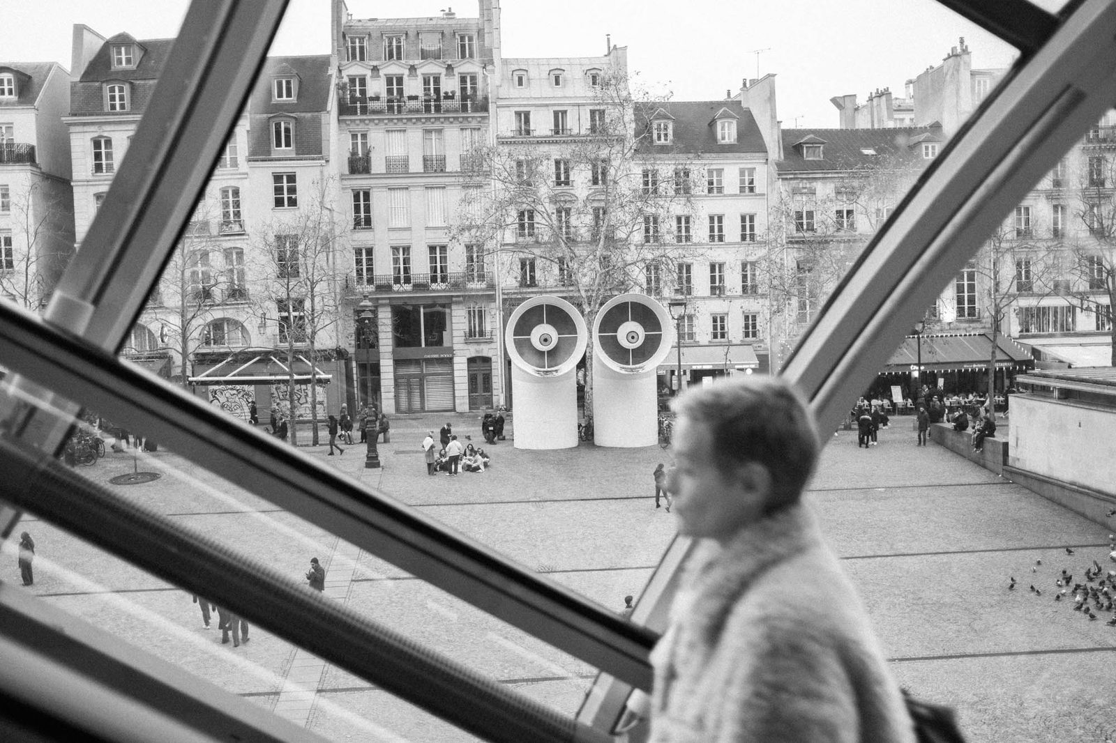 Black and white street photograph in Paris, a lone figure passing a Haussmann facade seen through a window frame.