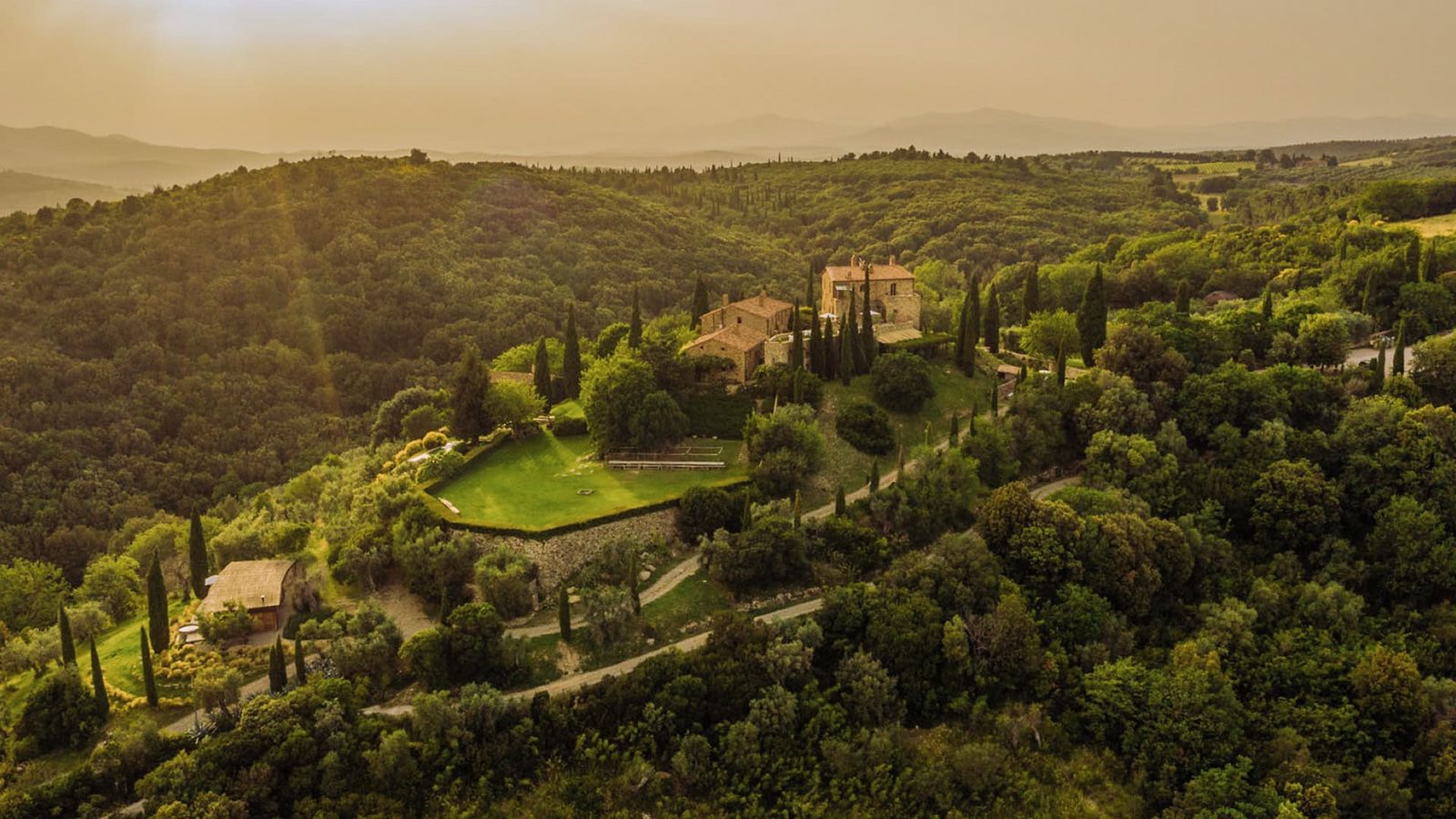 Aerial view of a Tuscan hilltop estate surrounded by woodland and cypress trees at golden hour.