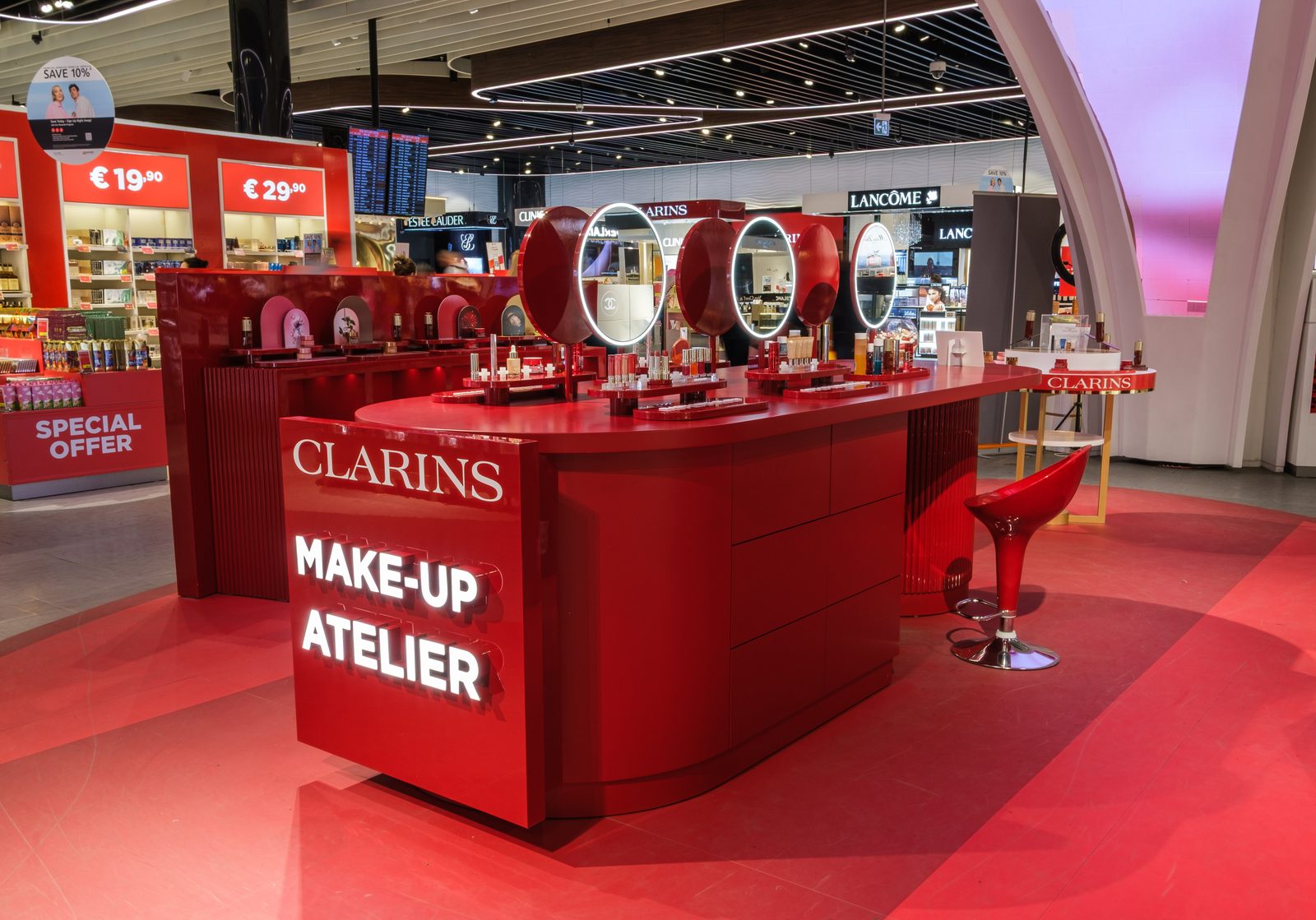 Clarins Make-Up Atelier counter with illuminated mirrors and stools on a red floor in an airport store.