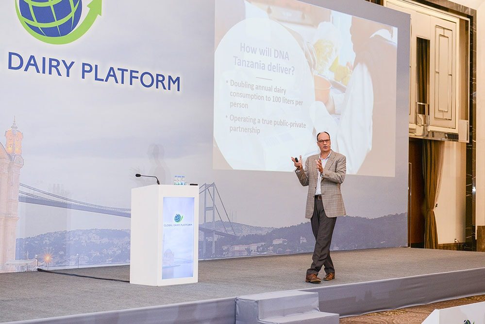 Speaker presenting on stage beside a branded lectern in front of a large projection screen.