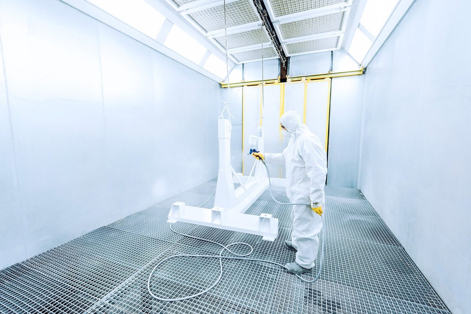 High-angle shot of a researcher in a cleanroom spraying equipment, wearing protective coveralls and gloves, working in a controlled environment with white walls and grid flooring.