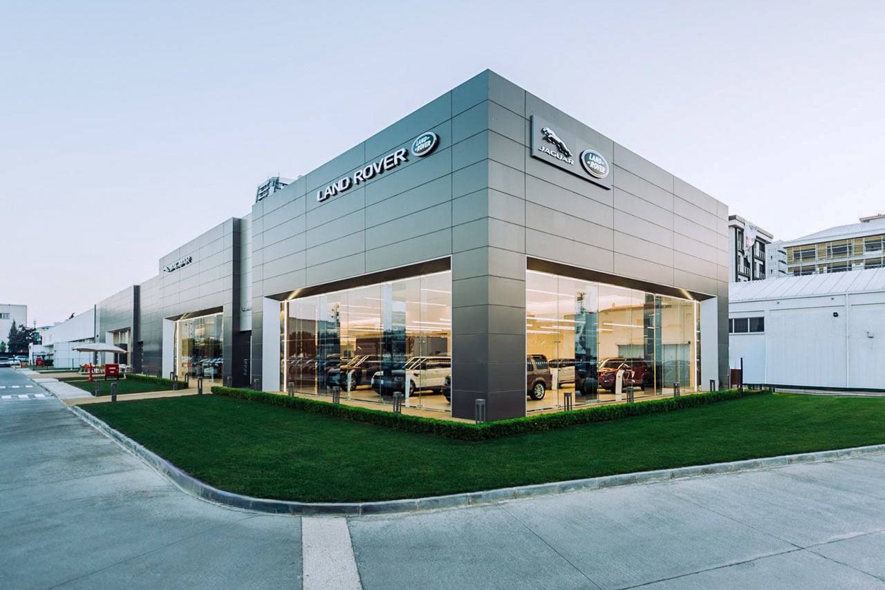 Vista de esquina de un concesionario de Jaguar y Land Rover, volumen acristalado de exposición sobre una explanada ajardinada.