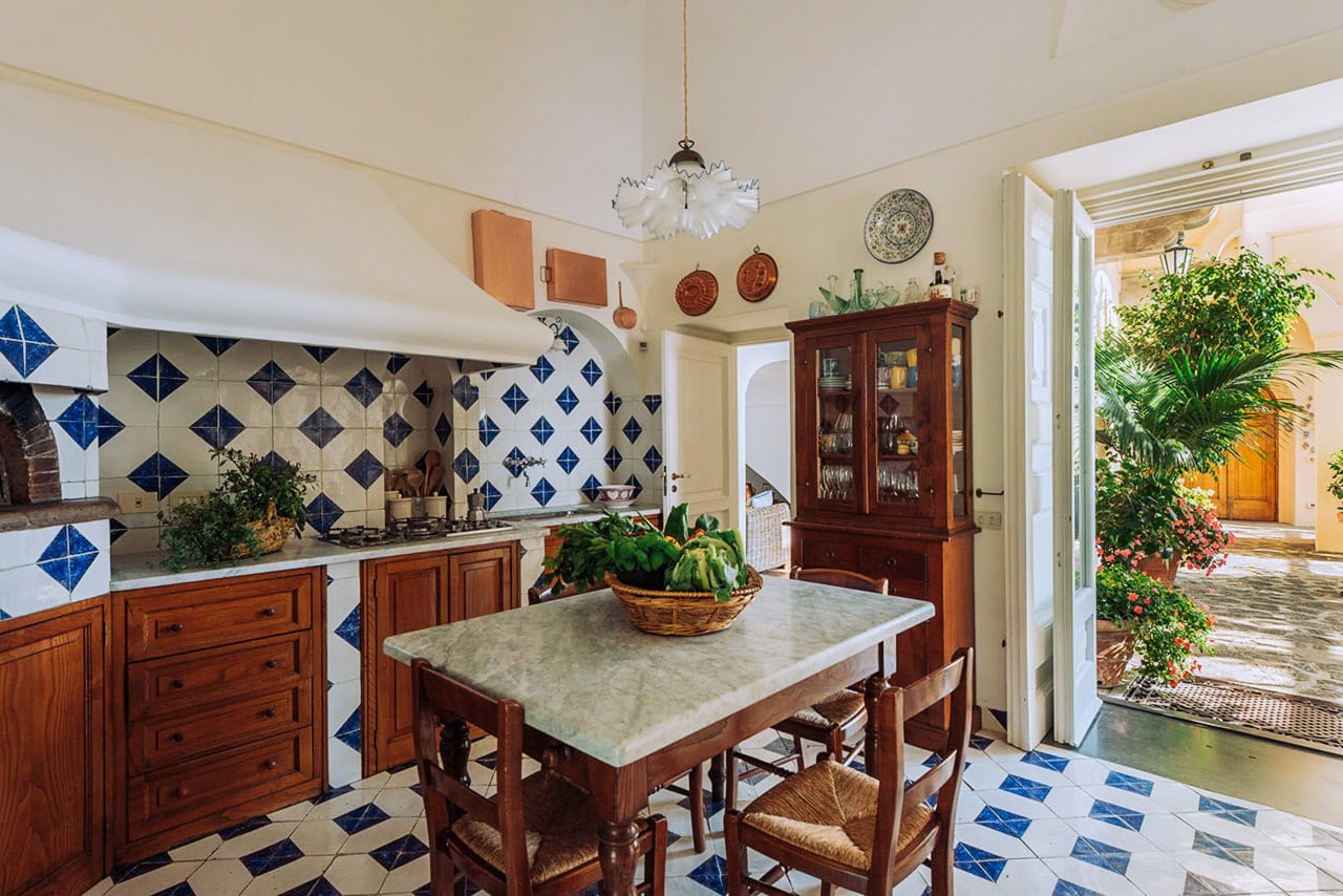 Farmhouse kitchen with wooden cabinets and blue tile backsplash, bright natural light, indoor-outdoor connection with potted plants, traditional decor, terrazzo flooring, cozy and inviting atmosphere.