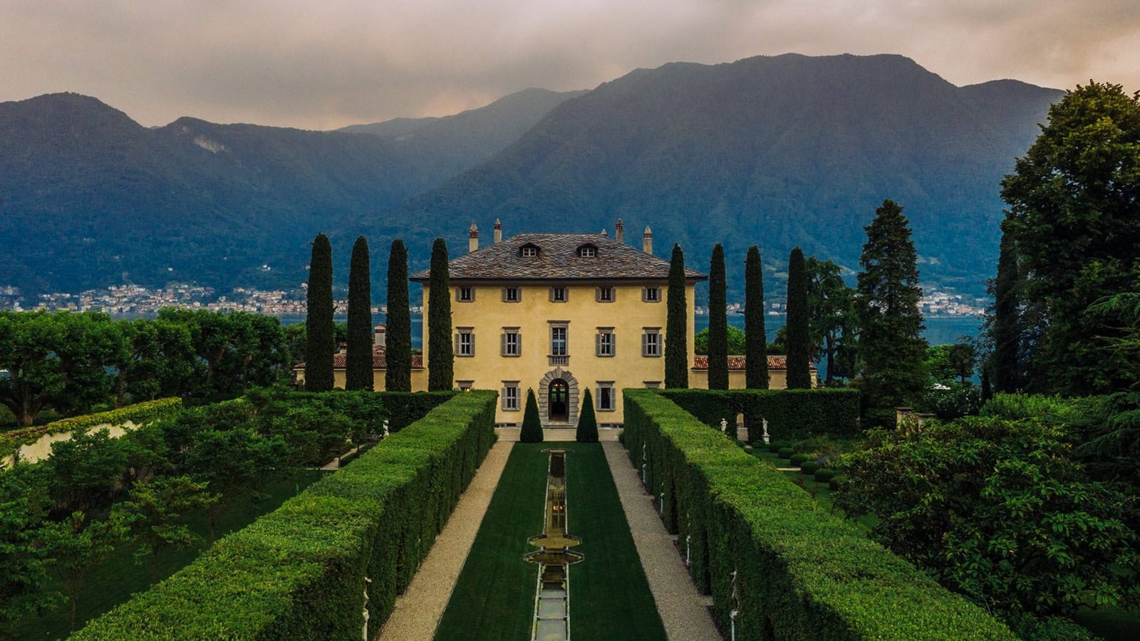 Lush Italian villa surrounded by manicured gardens and tall cypress trees with mountains in the background, captured in professional architectural photography. Photo shot by TKM Photo at Lake Como, Italy.