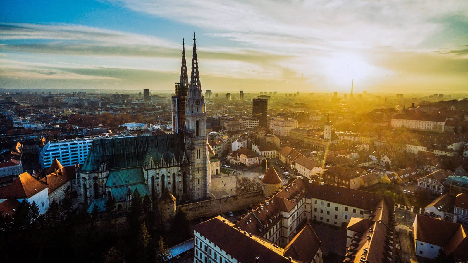 Aerial view of a historic European city at sunset, featuring a prominent Gothic cathedral with tall spires, surrounded by traditional buildings with red rooftops, bathed in golden light. Drone Photo of Zagreb Cathedral before the earthquake.