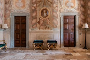 Ornate interior wall with fresco paintings, two wooden doors, and elegant decorative benches in a historic building in Madrid, Spain.
