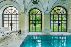 Luxurious indoor swimming pool with large arched windows and lush greenery outside, elegant architecture, natural light, serene ambiance, professional interior photography in Madrid, Spain, for hospitality and real estate purposes.
