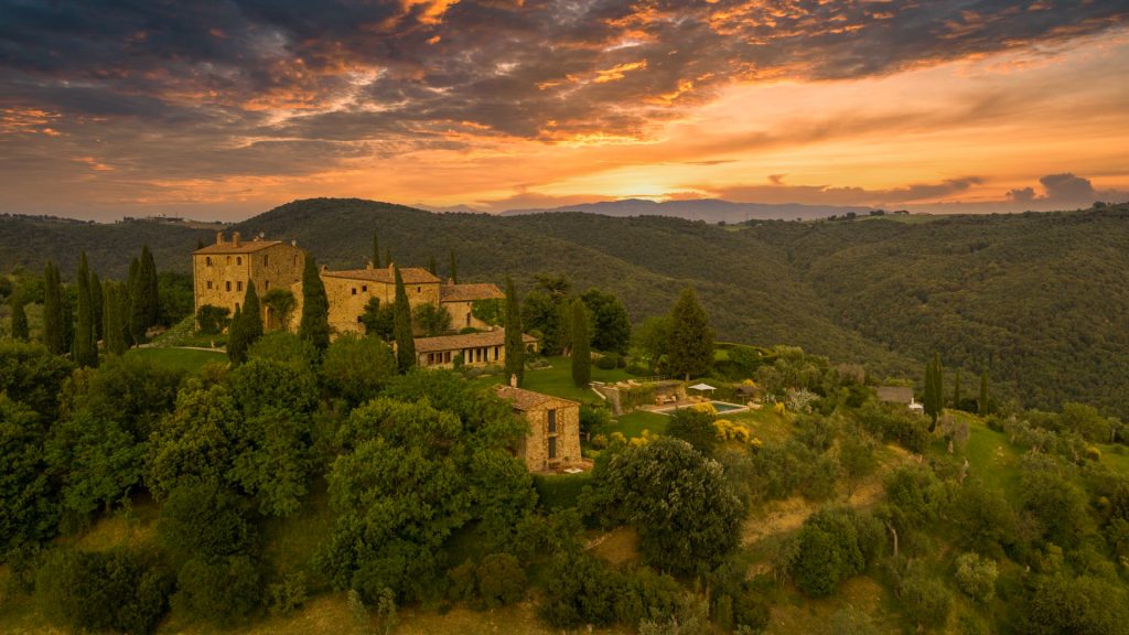 Breathtaking aerial view of a Tuscan-style hillside estate with iconic stone buildings, surrounded by lush green cypress trees during sunset. The vibrant sky creates a warm, inviting atmosphere, highlighting the scenic landscape.