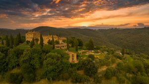 Breathtaking aerial view of a Tuscan-style hillside estate with iconic stone buildings, surrounded by lush green cypress trees during sunset. The vibrant sky creates a warm, inviting atmosphere, highlighting the scenic landscape.