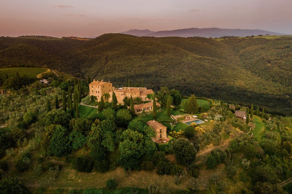 Lavish hillside estate surrounded by lush greenery and rolling hills in Tuscany at sunset. Aerial view showcasing the sprawling property, stone buildings, cypress trees, swimming pool, and picturesque landscape.