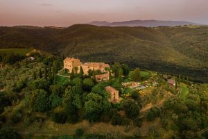 Lavish hillside estate surrounded by lush greenery and rolling hills in Tuscany at sunset. Aerial view showcasing the sprawling property, stone buildings, cypress trees, swimming pool, and picturesque landscape.
