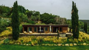 Secluded wooden cottage on a lush hillside surrounded by tall cypress trees and native plants, featuring a spacious deck with outdoor furniture and garden accents, offering a private retreat in nature.