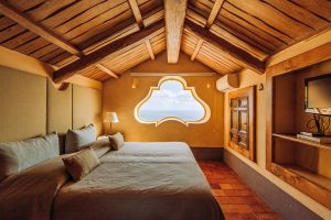 Cozy bedroom with a sloped wooden ceiling, a unique window shape offering ocean views, warm lighting, and rustic wooden accents, highlighting interior design for hospitality photography.