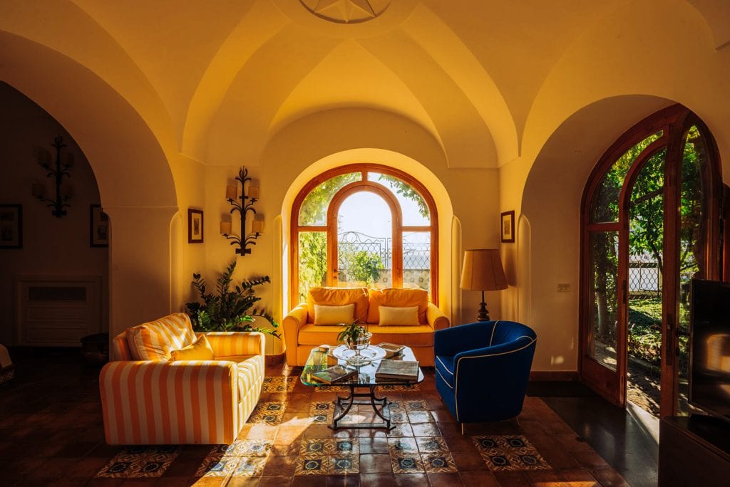 Colorful living room with arched windows, yellow, blue, and striped sofas, warm sunlight, and rustic tiled floor, showcasing interior design photography by TKM Photo in Madrid.