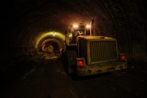 Excavator working inside an underground tunnel at night, construction equipment on site, industrial underground project, heavy machinery in a mining or tunneling environment, dark and gritty atmosphere with artificial lighting.
