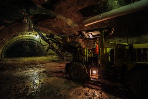 Underground mining tunnel with heavy machinery and a worker in safety gear illuminated by artificial lighting indoors.