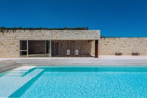 Modern residential building with a swimming pool, stone wall exterior, and a clear blue sky, showcasing architectural and outdoor lifestyle photography by TKM Photo in Madrid.