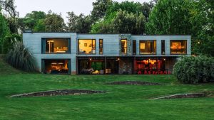 Modern architectural house with large glass windows and a lush green lawn, illuminated from within, surrounded by trees and landscape, captured in professional photography.