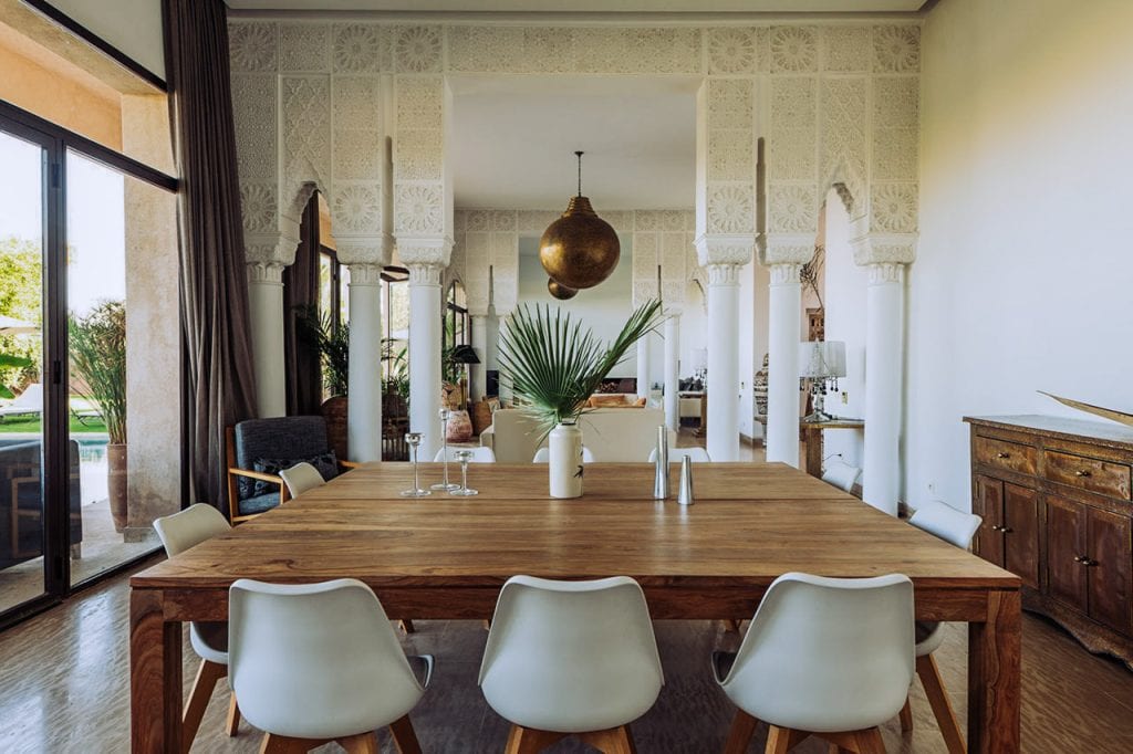 Modern dining room with wooden table, white chairs, decorative columns, and large sliding glass door overlooking outdoor garden, well-lit interior with stylish decor and lush greenery outside.