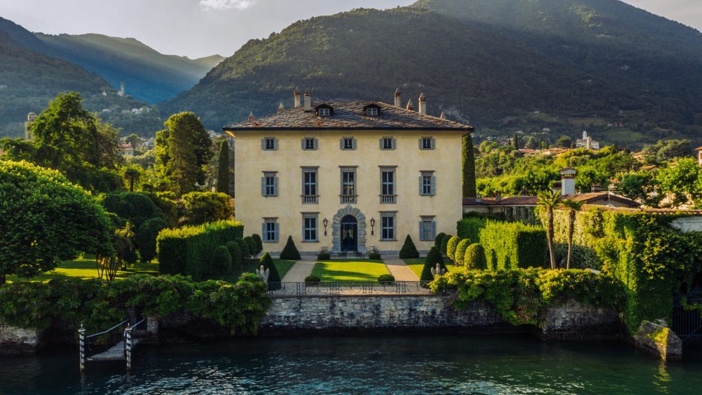 Lush Italian villa surrounded by manicured gardens and tall cypress trees with mountains in the background, captured in professional architectural photography. Photo shot by TKM Photo at Lake Como, Italy.