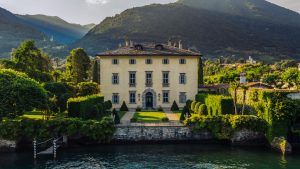 Lush Italian villa surrounded by manicured gardens and tall cypress trees with mountains in the background, captured in professional architectural photography. Photo shot by TKM Photo at Lake Como, Italy.