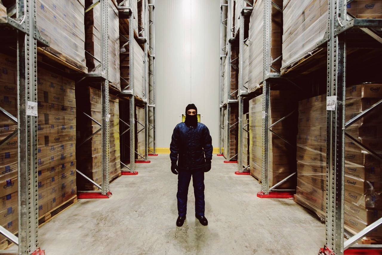 1. A person wearing a black mask and dark clothing standing in a warehouse aisle with tall shelves filled with boxes on either side.