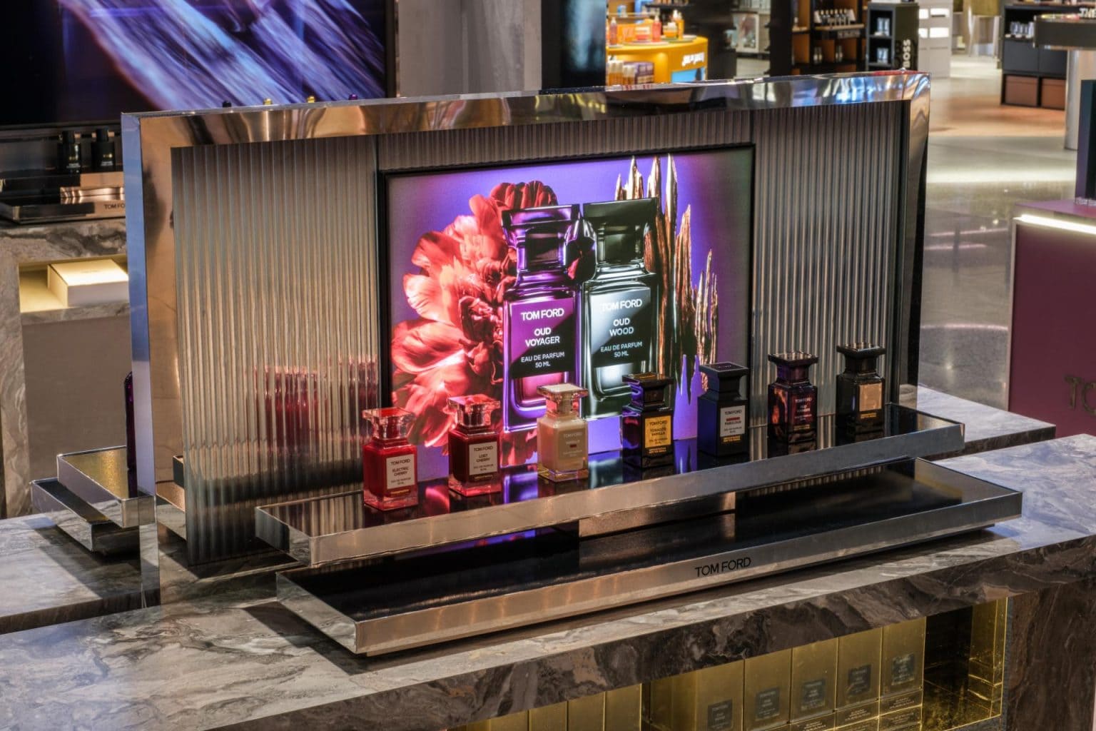 A retail perfume display featuring Tom Ford fragrances with colorful packaging and modern design, showcased on a sleek metallic stand in a luxury store setting.